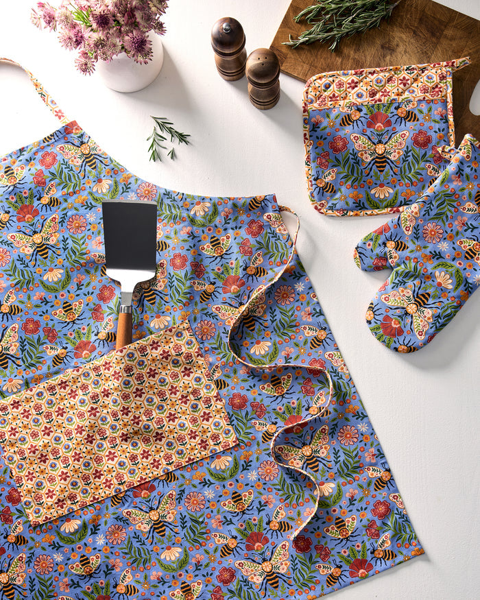Bee Tapestry blue floral-and-bee print apron displayed with its neck strap and front pocket among coordinating kitchen textiles.