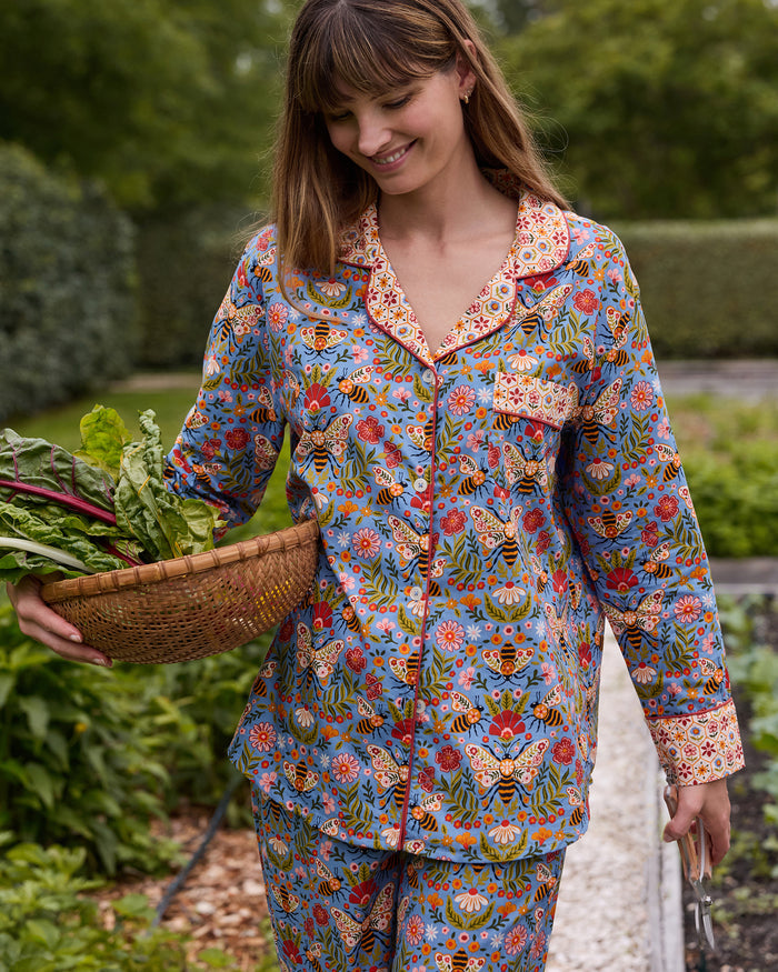 Person wearing the Delphinium Blue Bee Tapestry long pajama set and carrying a woven basket of leafy vegetables in a garden.