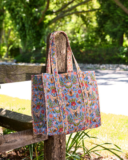 Bee Tapestry traveler tote hanging from a rustic wooden fence, shown at an angle with its blue bee-and-floral print.