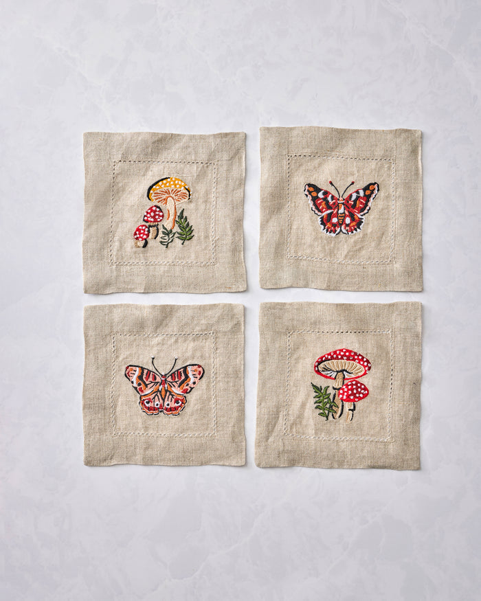 Four square embroidered cocktail napkins displayed in a two-by-two arrangement, with mushroom and butterfly designs.