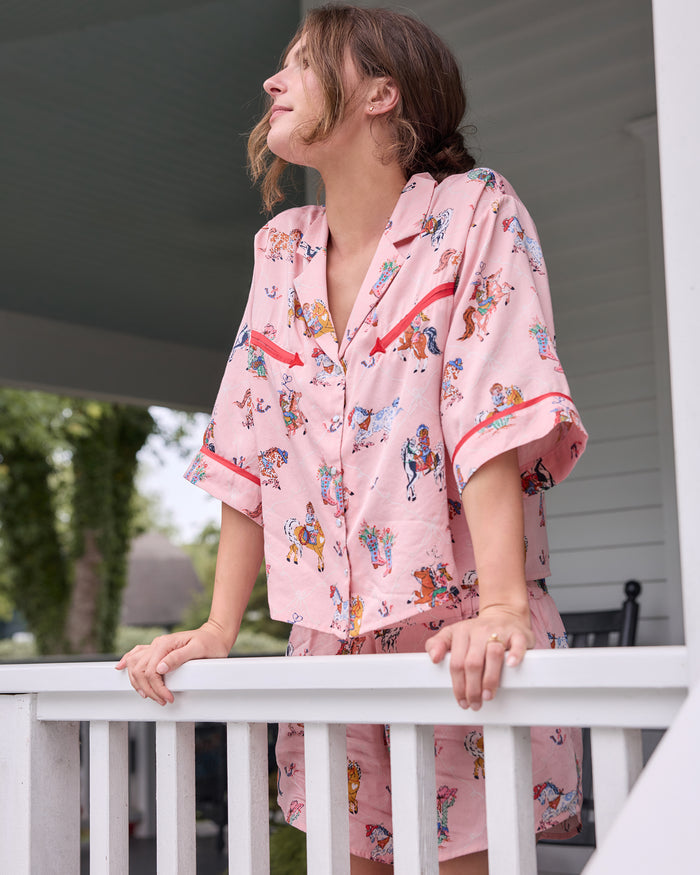 Three-quarter view of a short-sleeve pajama top and matching shorts with a colorful cowgirl, horse, and boot print, posed at a porch railing.
