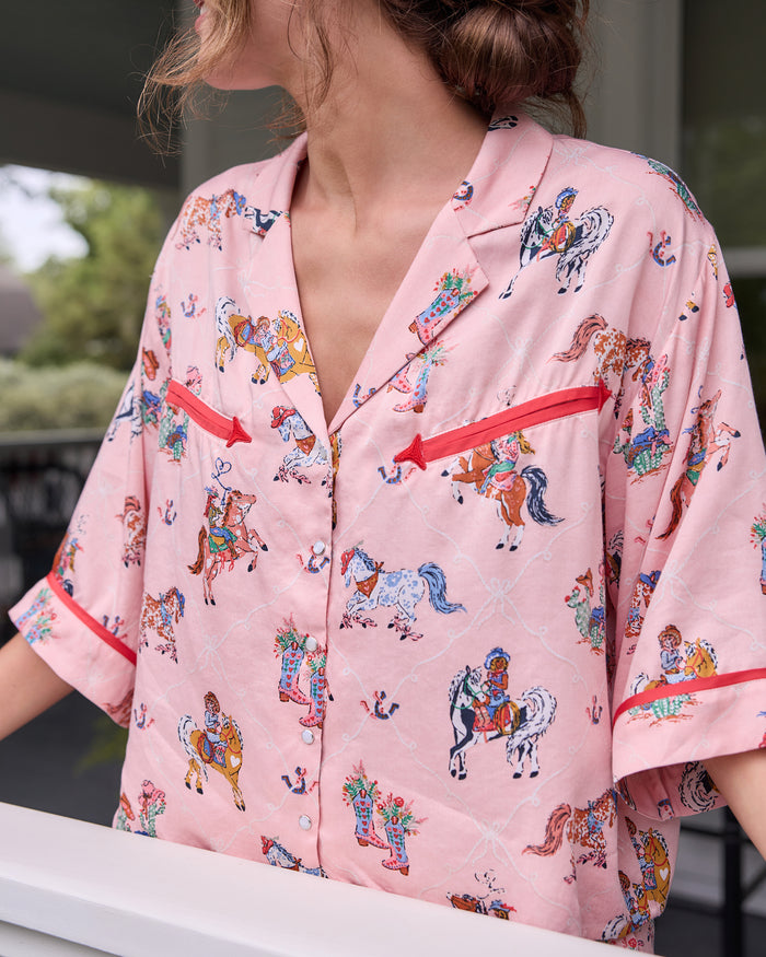 Close-up of the cropped pink pajama top with a horse-and-cowgirl print, red trim, notched collar, and front buttons.
